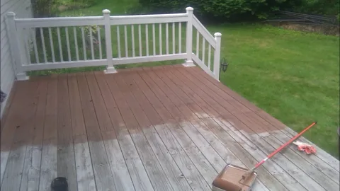 Deck resurfacing services in progress on a weathered wooden deck partly coated in reddish-brown stain, with a long-handled brush and tray beside a green lawn and white railing.