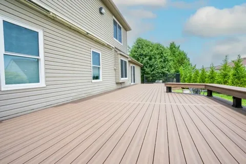 Deck resurfacing services restored this spacious, sunlit wood deck along a beige-sided house, with a low railing, green trees, and blue sky.